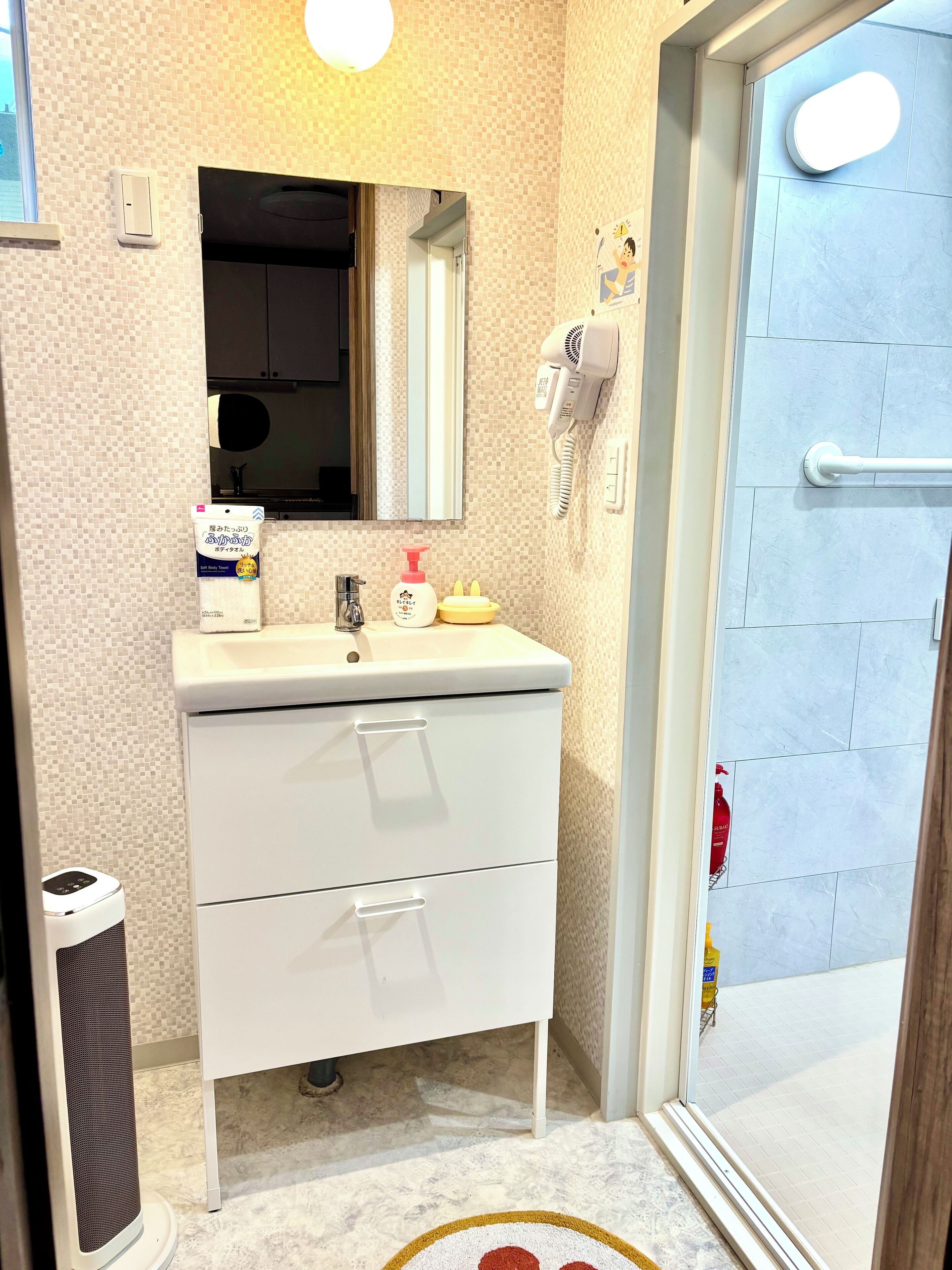 Ein Badezimmer mit einem weißen Waschbecken, einem Spiegel und einem an der Wand befestigten Föhn. Es gibt eine weiße Kommode mit Schubladen und ein weißes Luftreinigergerät.