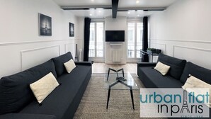 Un salon moderne avec deux canapés noirs, une table basse en verre et un téléviseur à écran plat fixé au mur.
