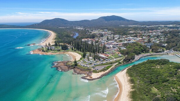 An aerial view of a coastal town with a beach, clear blue waters, and a mix of residential and commercial buildings.