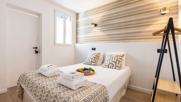 A neatly made bed with a patterned bedspread, a tray of fruits and a bottle of water, and a wooden shelf unit to the right. - Pleasant Studio - 1P - Beaugrenelle / Tour Eiffel (Paris)