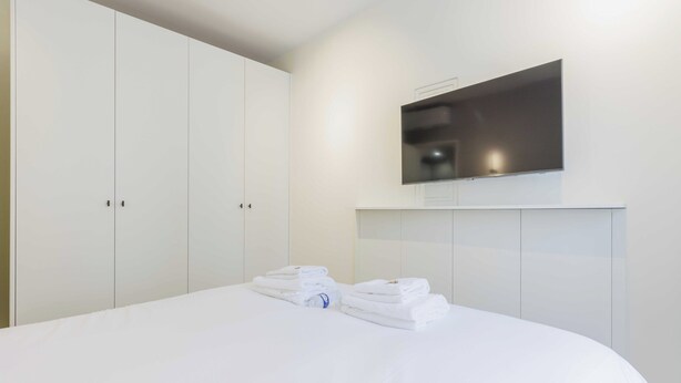 A hotel room with a large bed, a flat-screen TV mounted on the wall, and a wardrobe with multiple doors. - Cozy Apartment 2br/6p - Sacre-coeur / Montmartre (Paris)