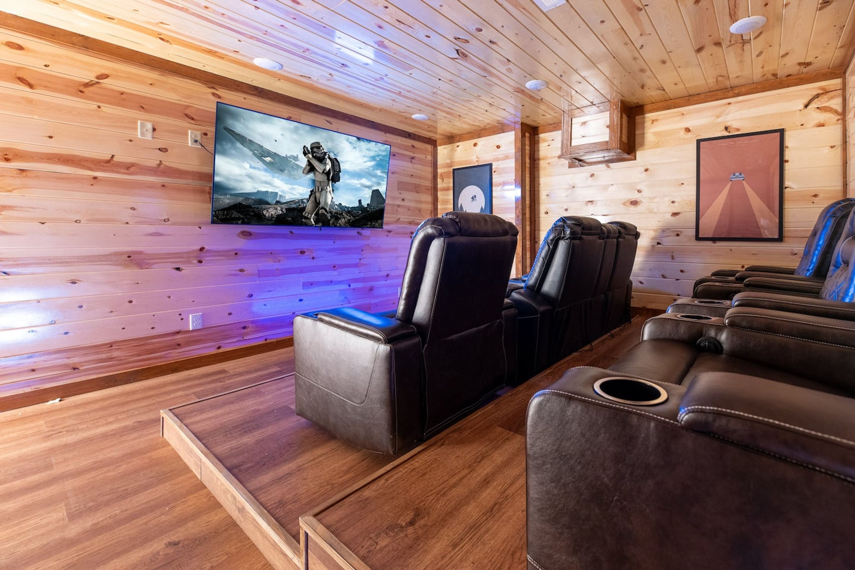 A wooden-paneled room with a large flat-screen TV mounted on the wall, leather reclining chairs, and a wooden coffee table.