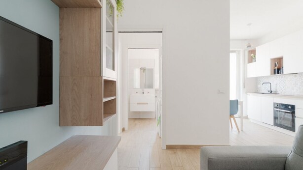 A modern living room with a flat-screen TV, wooden shelving, a grey sofa, and a kitchen in the background. - GuestHost - Naviglio Grande Bright Apartment (Milan)