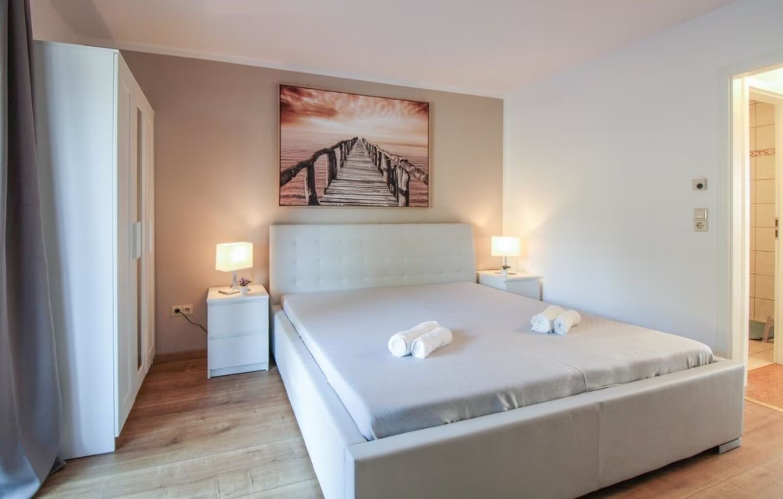 Ein ordentlich eingerichtetes Schlafzimmer mit einem großen Bett, zwei Nachttischen mit Lampen, einem Bild an der Wand und Holzböden.