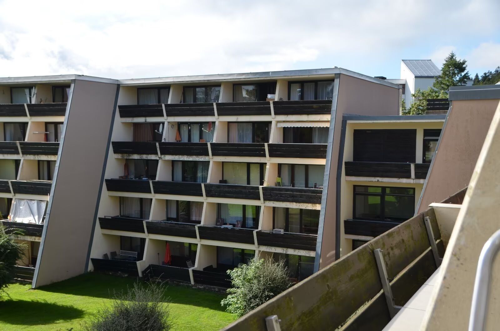 A multi-story building with balconies, a flat roof, and a small garden area.