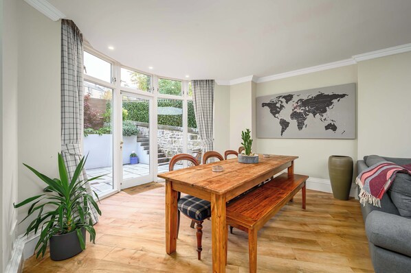 A dining area with a wooden table, chairs, and a bench. There is a potted plant, a world map on the wall, and a grey sofa.
