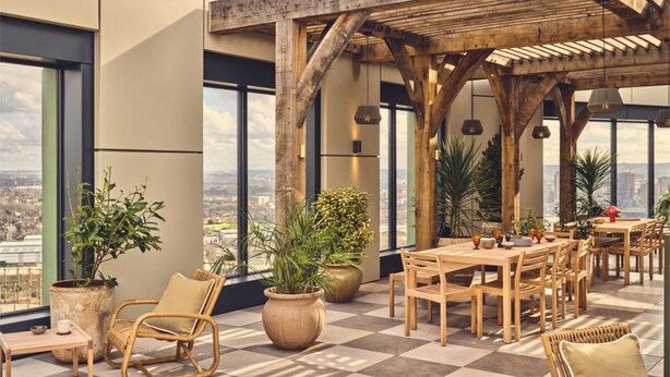 A rooftop terrace with wooden furniture, large potted plants, and a city view.