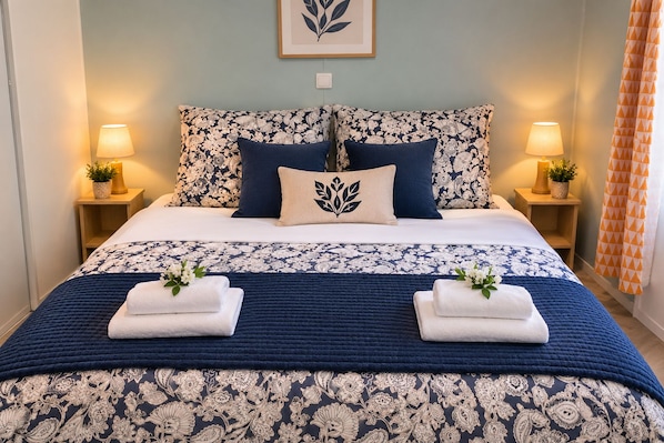 Uma cama bem arrumada com colcha e almofadas estampadas com flores, duas mesas de cabeceira com abajures e uma obra de arte emoldurada na parede.