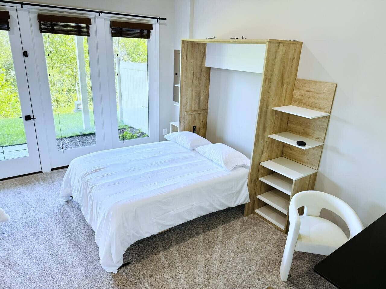 A bedroom with a bed, a white chair, a wooden shelf unit, and a sliding glass door leading to a garden.