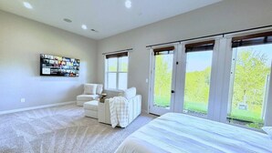 A bedroom with a large bed, a white armchair, and a sofa. There are two windows with blinds and a door leading to a balcony.