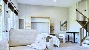 A modern living room with a sofa, a small table, a staircase, and a bed in the background.