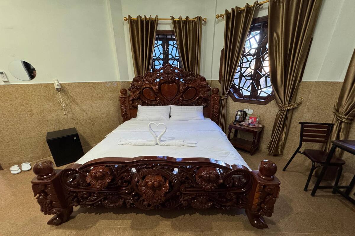 A wooden bed with intricate carvings, a heart-shaped towel, and a small bedside table with a kettle.