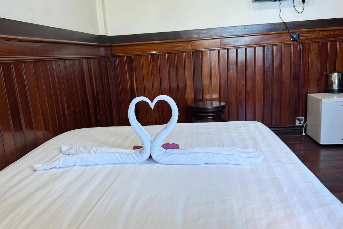 A neatly made bed with a towel swan centerpiece, a wooden headboard, a small bedside table, and a TV mounted on the wall.