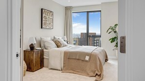 A modern bedroom with a large window, a bedside table with a lamp, and a bed with a beige cover. - Stylish One-bedroom Apartment Close to Stratford (London)