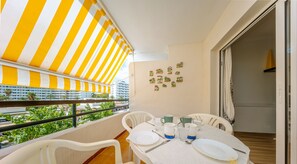 Un balcón con un toldo a rayas, sillas blancas, una mesa dispuesta con platos, vasos y una cuchara.