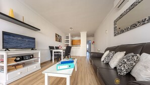 A modern living room with a flat-screen TV, a white coffee table, a brown leather sofa, and a dining area in the background.