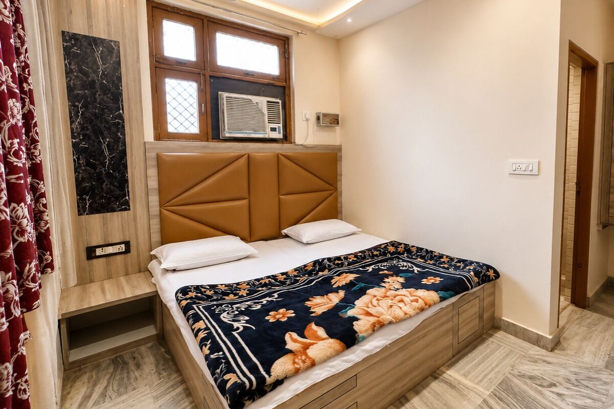 A bedroom with a bed, a headboard, a window with an air conditioner, a wardrobe, and a wall-mounted outlet.