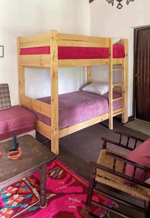 A bunk bed with a wooden frame and purple bedding, a small table with a basket, a colorful rug, and a wooden chair.