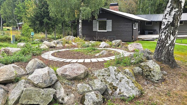 A cabin with a stone pathway leading to a circular seating area, surrounded by rocks and trees.