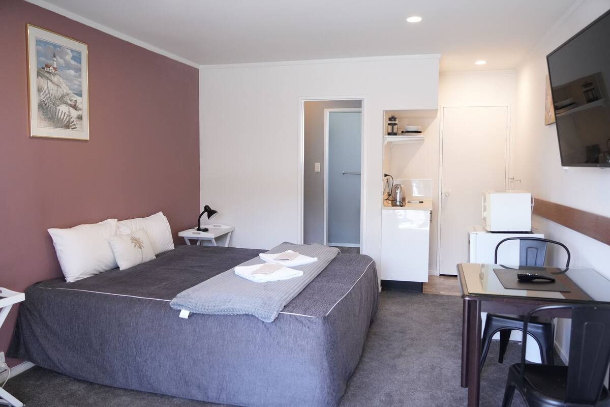 A hotel room with a bed, a desk, a chair, a television, and a kitchenette.