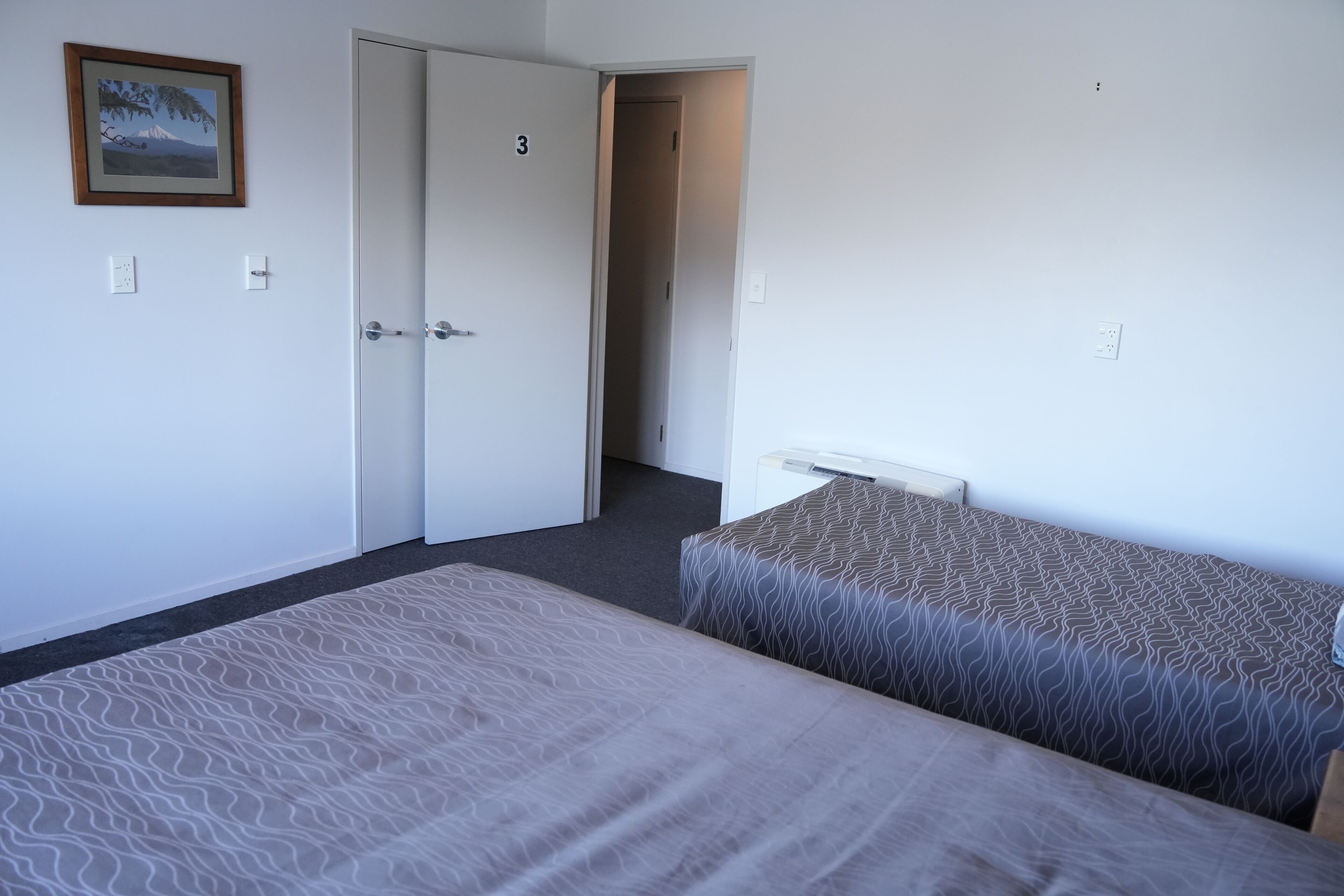 A room with two beds, a door with a number, a framed picture on the wall, and a small table.