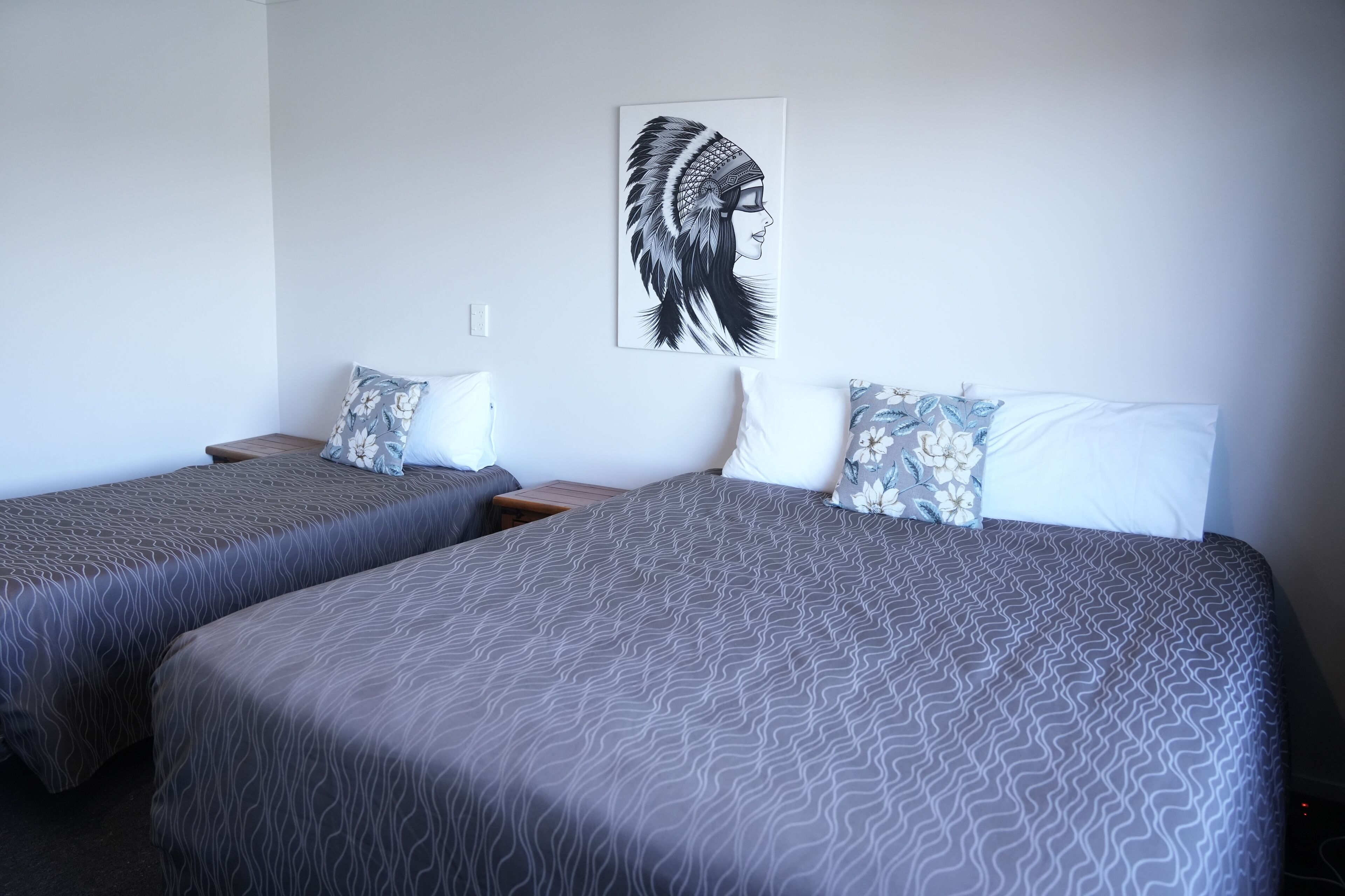 A hotel room with two single beds, each with a patterned bedspread and matching pillows. A framed artwork is displayed on the wall above the beds.