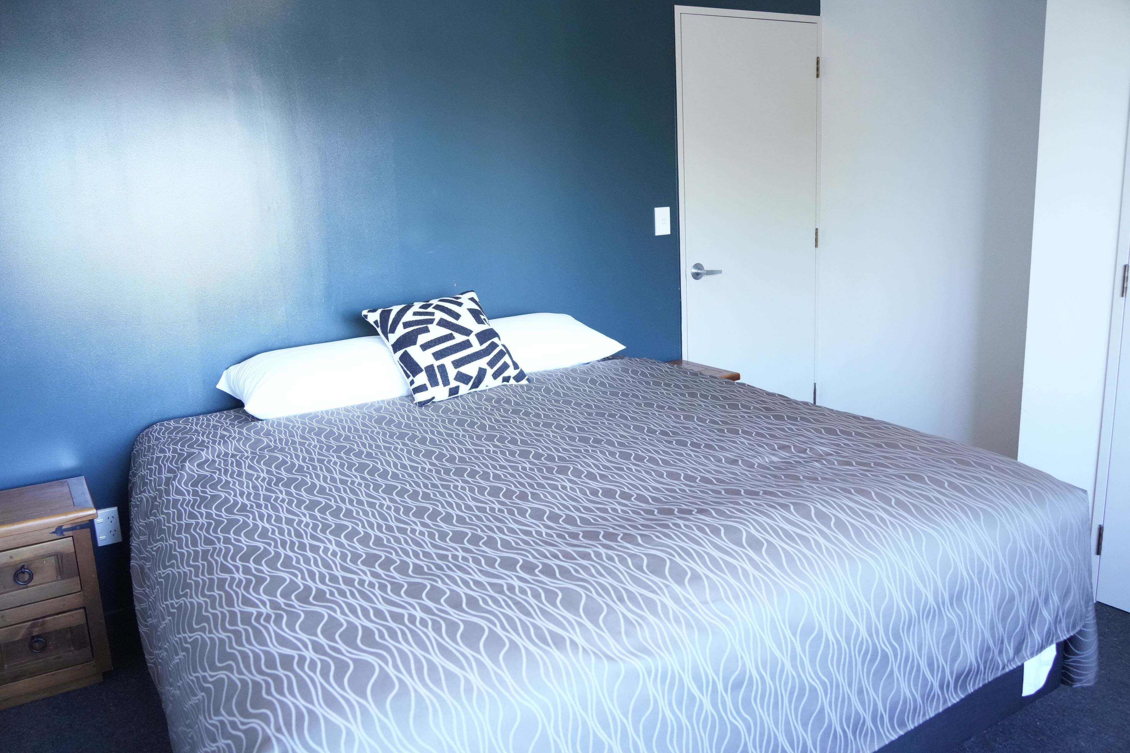 Un letto rifatto con una coperta a fantasia, un comodino in legno con cassetti e una porta bianca in una stanza dalle pareti blu.