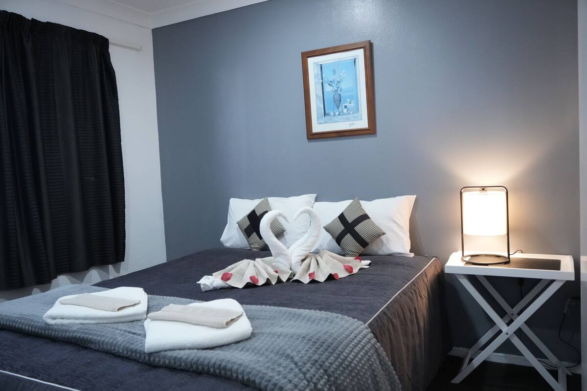 A neatly made bed with a towel and a decorative swan arrangement, a framed picture on the wall, and a bedside table with a lamp.