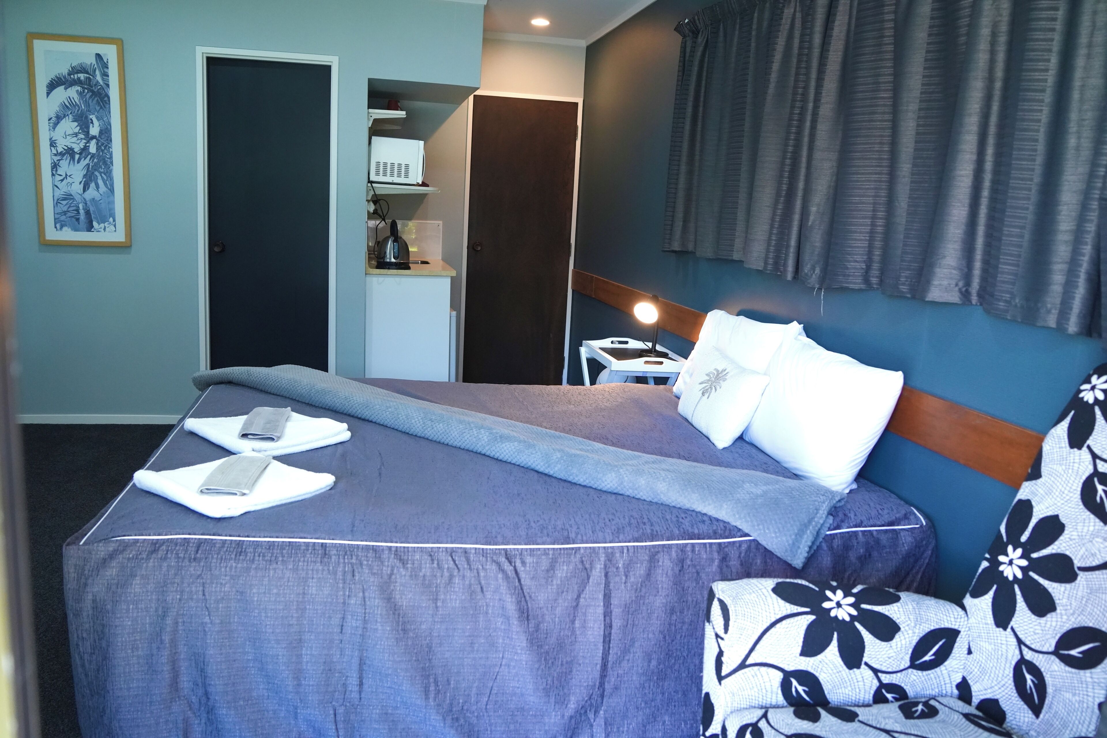 Un lit bien fait, recouvert d’une couvre-lit bleue, avec un oreiller blanc et un coussin décoratif. Une table de chevet, sur laquelle se trouvent une lampe et un petit objet blanc, est placée à côté du lit. La pièce a un mur bleu et une fenêtre avec des rideaux.