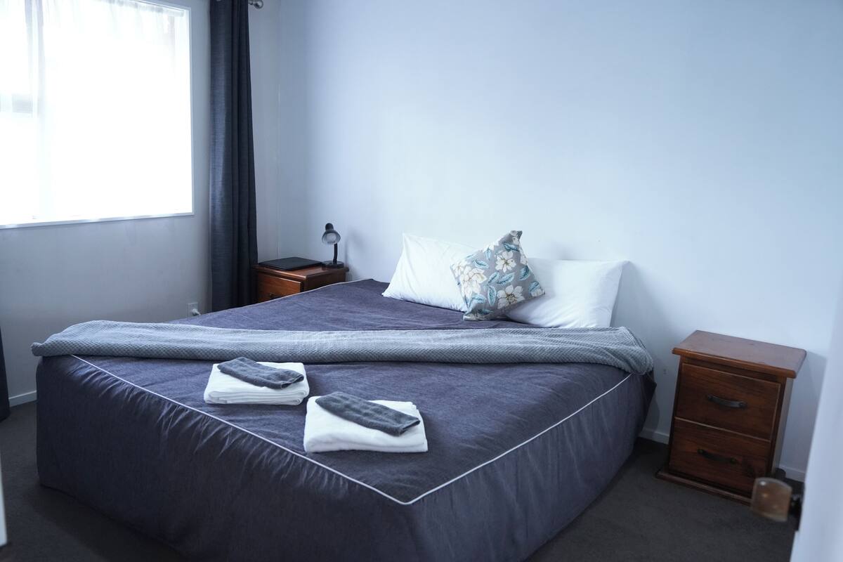 A neatly made bed with a dark blue comforter and pillows, a small bedside table with a lamp, and a nightstand with a drawer.
