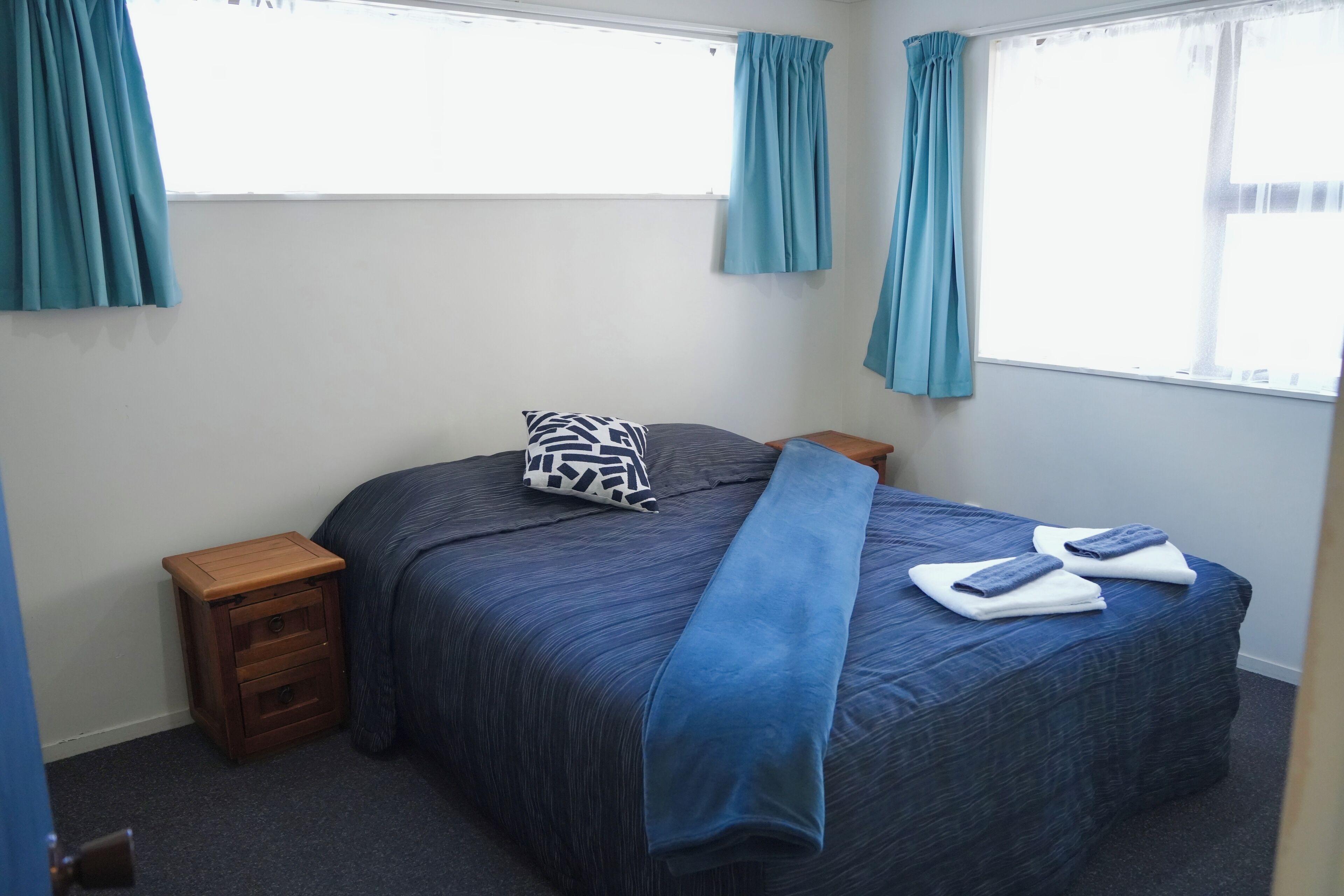 A neatly made bed with a blue bedspread, a patterned pillow, and two folded towels. There is a wooden nightstand beside the bed.