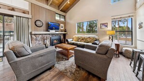A cozy living room with a stone fireplace, a television, and a large window offering a view of trees.