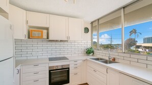 Cocina moderna con armarios blancos, horno empotrado, microondas y un amplio ventanal con vistas a un paisaje tropical.