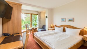 Ein Hotelzimmer mit einem Bett, einem Schreibtisch mit Telefon, einem Sessel, einem kleinen Tisch, einem Fernseher und einem Fenster mit Blick ins Grüne.