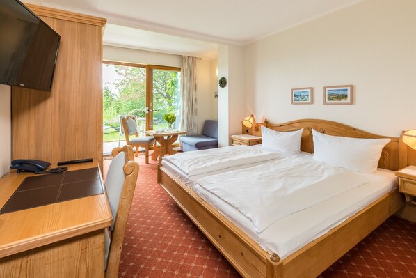 Ein Hotelzimmer mit einem Bett, einem Schreibtisch mit Telefon, einem Sessel, einem kleinen Tisch, einem Fernseher und einem Fenster mit Blick ins Grüne.