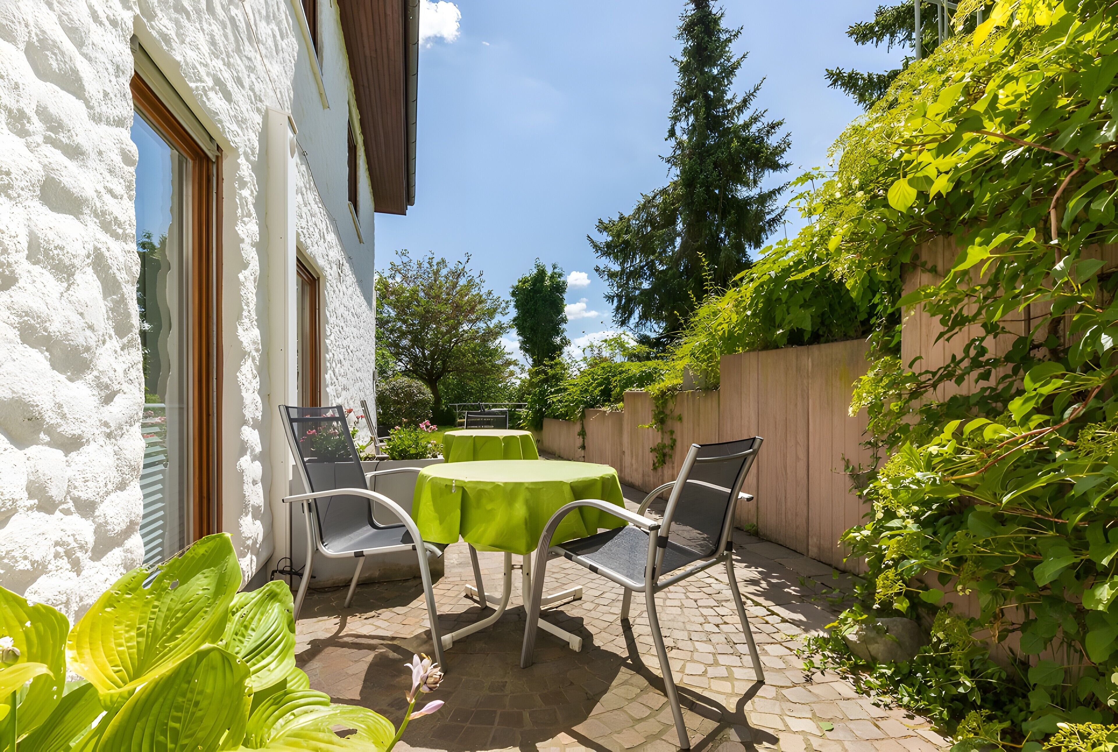 A patio with a table and chairs, a white wall, and greenery.