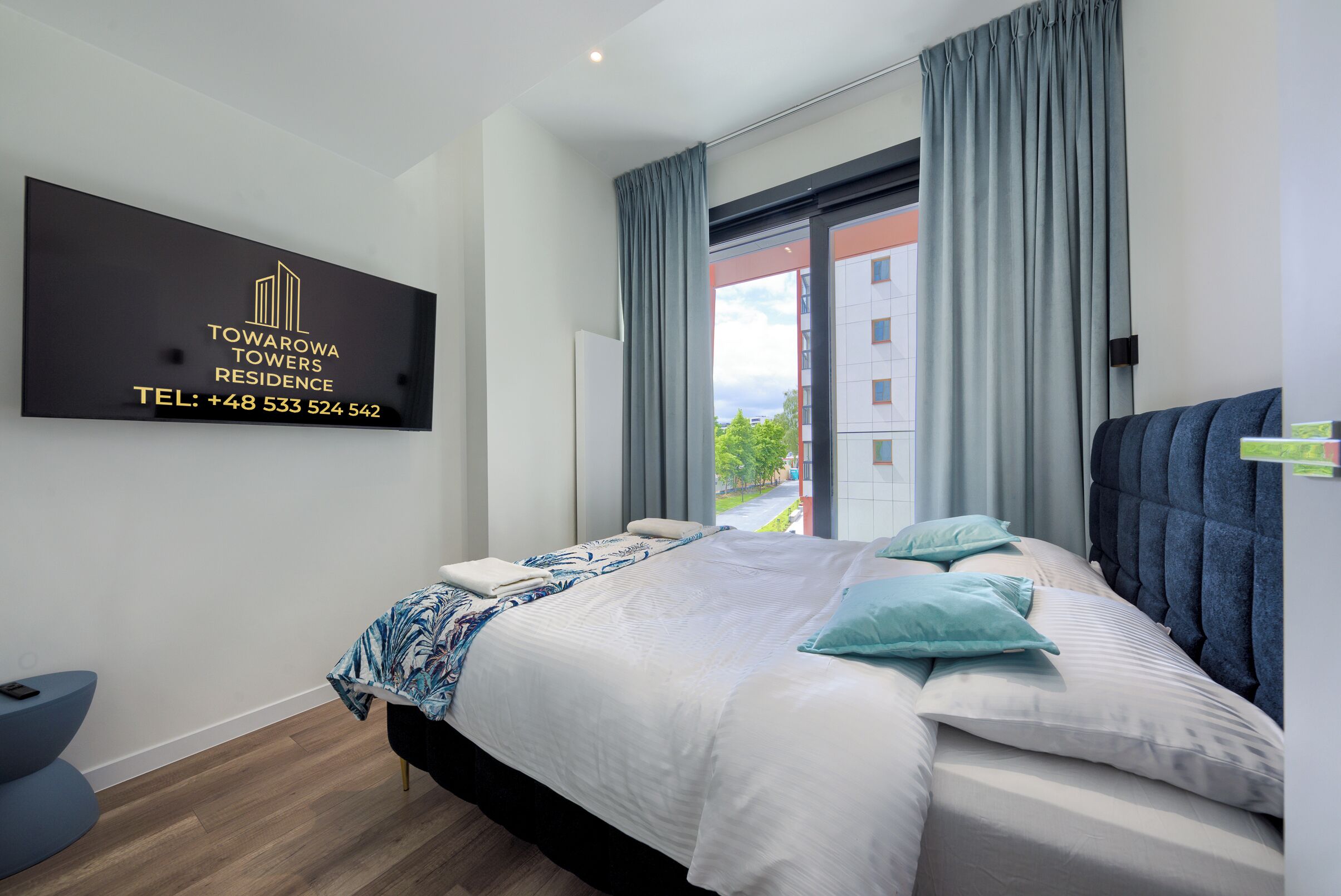 A modern hotel room with a bed, a view of the outside, and a sign on the wall with the hotel's name and contact number.