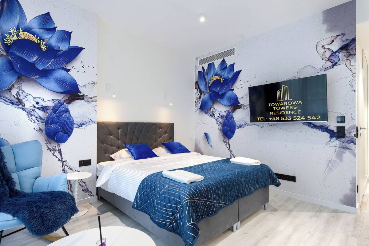 Ein modernes Schlafzimmer mit einem großen Bett, einem blauen Sessel und einem Fernseher, der ein Logo und Kontaktinformationen zeigt.