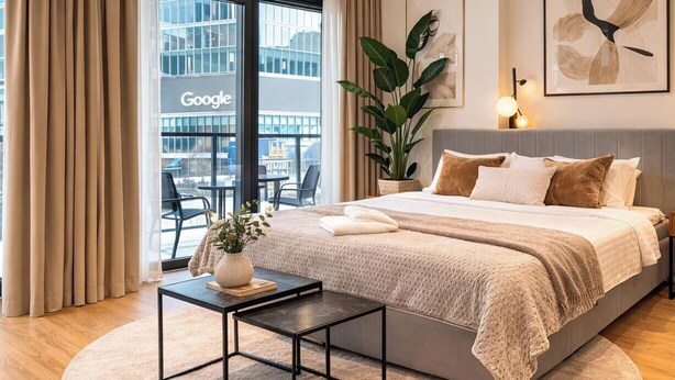 A modern hotel room with a large bed, a small table, a potted plant, and a view of a building with a Google sign. - Towarowa Towers Residences (Warsaw)