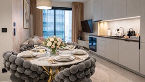A modern dining room with a round table set for six, a large window with curtains, a flat-screen TV, and a kitchen area with a built-in oven and microwave. - Towarowa Towers Residences (Warsaw)