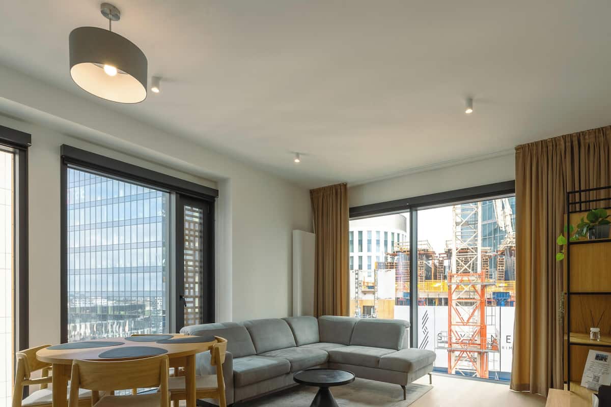 Ein modernes Wohnzimmer mit einer Eckcouch, einem Esstisch mit Stühlen, großen Fenstern und Blick auf die Stadt sowie einem Bücherregal.