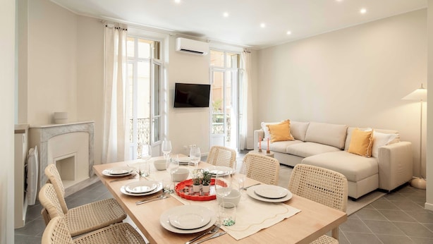 Un comedor con una mesa preparada para cuatro personas, rodeado de sillas de mimbre, un sofá y un televisor fijado a la pared.