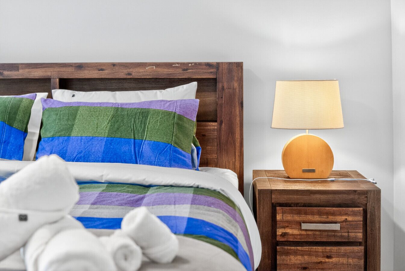 Een bed met gestreepte lakens, een houten nachtkastje met een lamp en een houten hoofdbord.