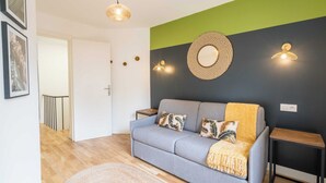 Un salon moderne avec un canapé gris, deux coussins, une décoration murale tissée et une petite table d’appoint.