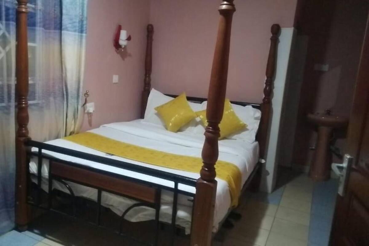 Uma cama de dossel com colcha e almofadas amarelas, cabeceira de madeira e cortina no lado esquerdo.