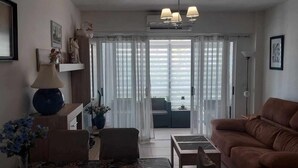 A living room with a sofa, a coffee table, a lamp, and a vase with flowers. There is a window with curtains and a view of the outside. - Apartamento Beata Para 4 Personas (Calpe)
