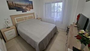 A neatly arranged bedroom with a bed, bedside tables, a desk with a computer, and a window with sheer curtains. - Apartamento Beata Para 4 Personas (Calpe)