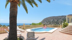 Zona de piscina con vista al mar, una palmera y una montaña al fondo.