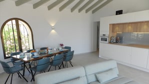 Una cocina moderna con armarios blancos y una encimera de madera, un comedor con mesa de vidrio y sillas azules, y un sofá de color gris claro.