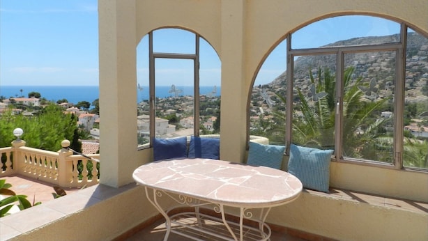 Un balcón con mesa y sillas, ventanas amplias con vistas a un pueblo costero y colinas.